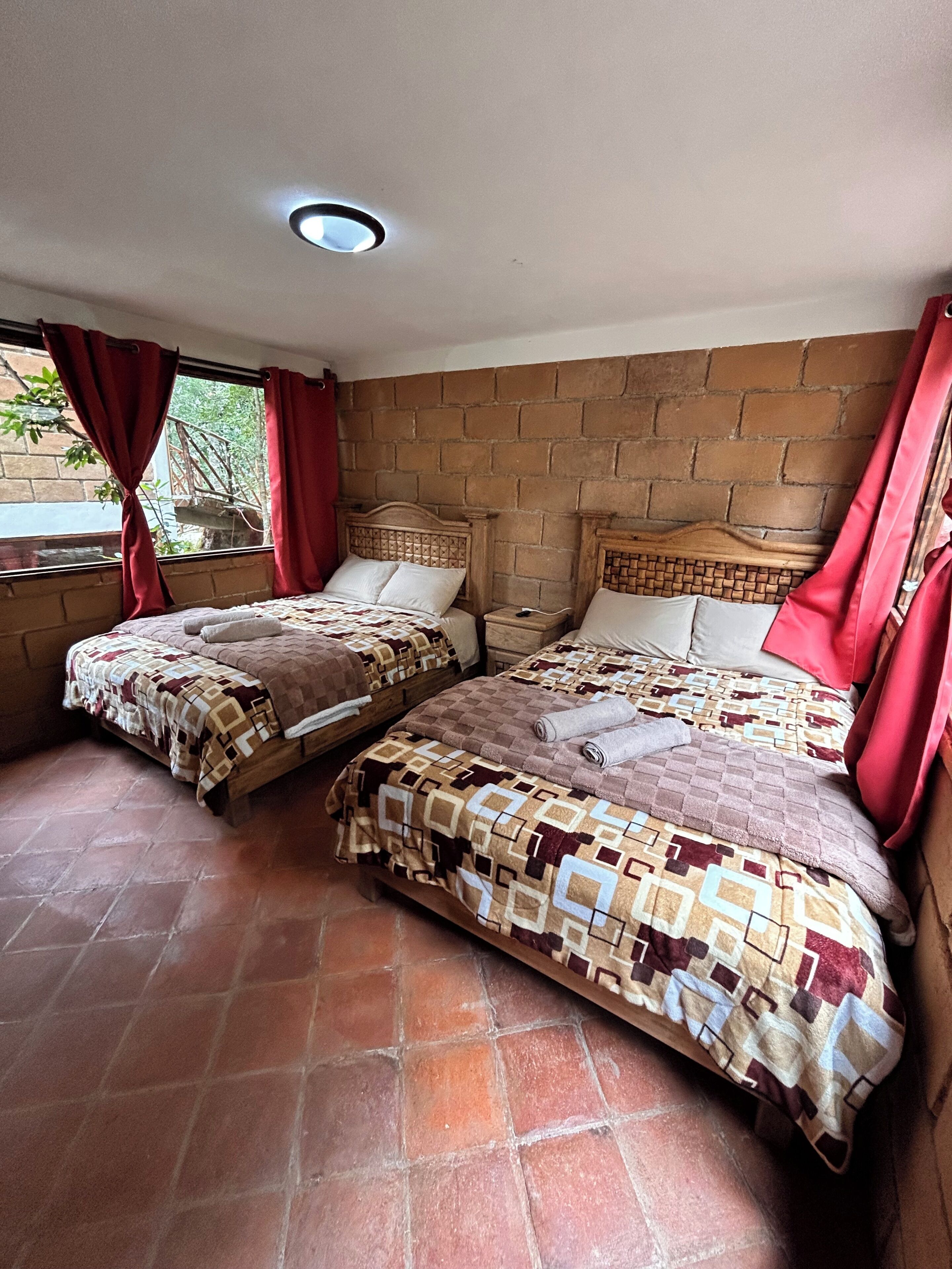 A room with two beds, a brick wall, and a window with red curtains.
