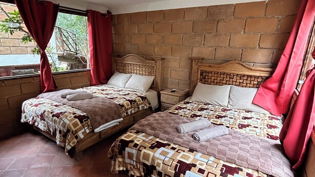 Una habitación con dos camas, un muro de ladrillo y una ventana con cortinas rojas.