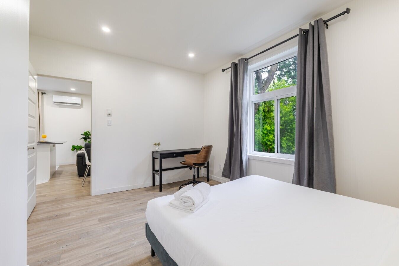 A modern hotel room with a bed, a desk, and a chair. There is a window with curtains, and a view of greenery outside.