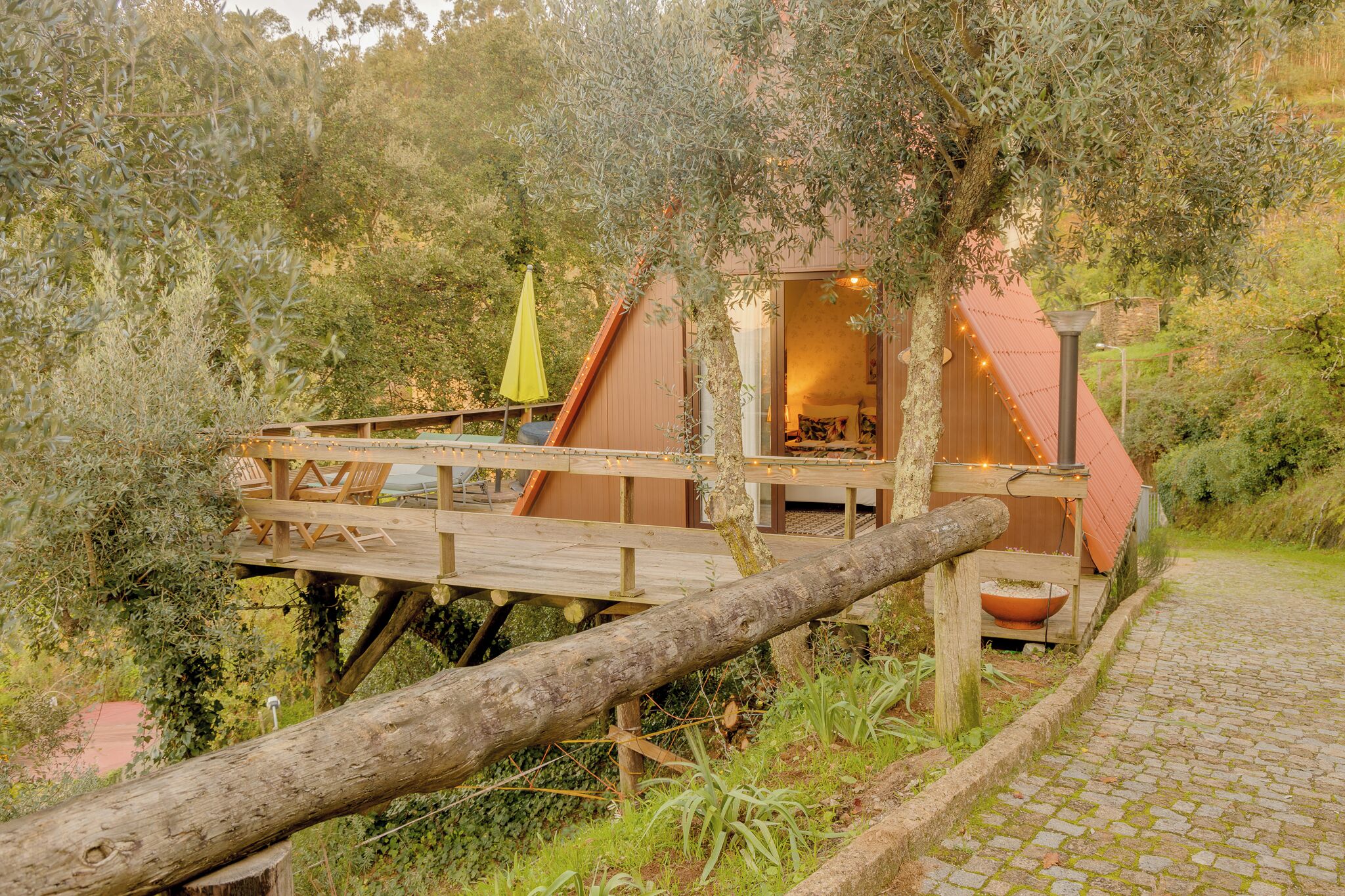 Una terraza de madera con sillas y una mesa, un pequeño edificio de techo rojo, y un sendero que lo conduce.