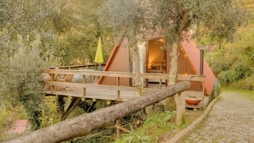 Una terraza de madera con sillas y una mesa, un pequeño edificio de techo rojo, y un sendero que lo conduce.