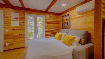 A room with wooden walls, a bed with white bedding and yellow pillows, a grey sofa, and a window with a view of trees.