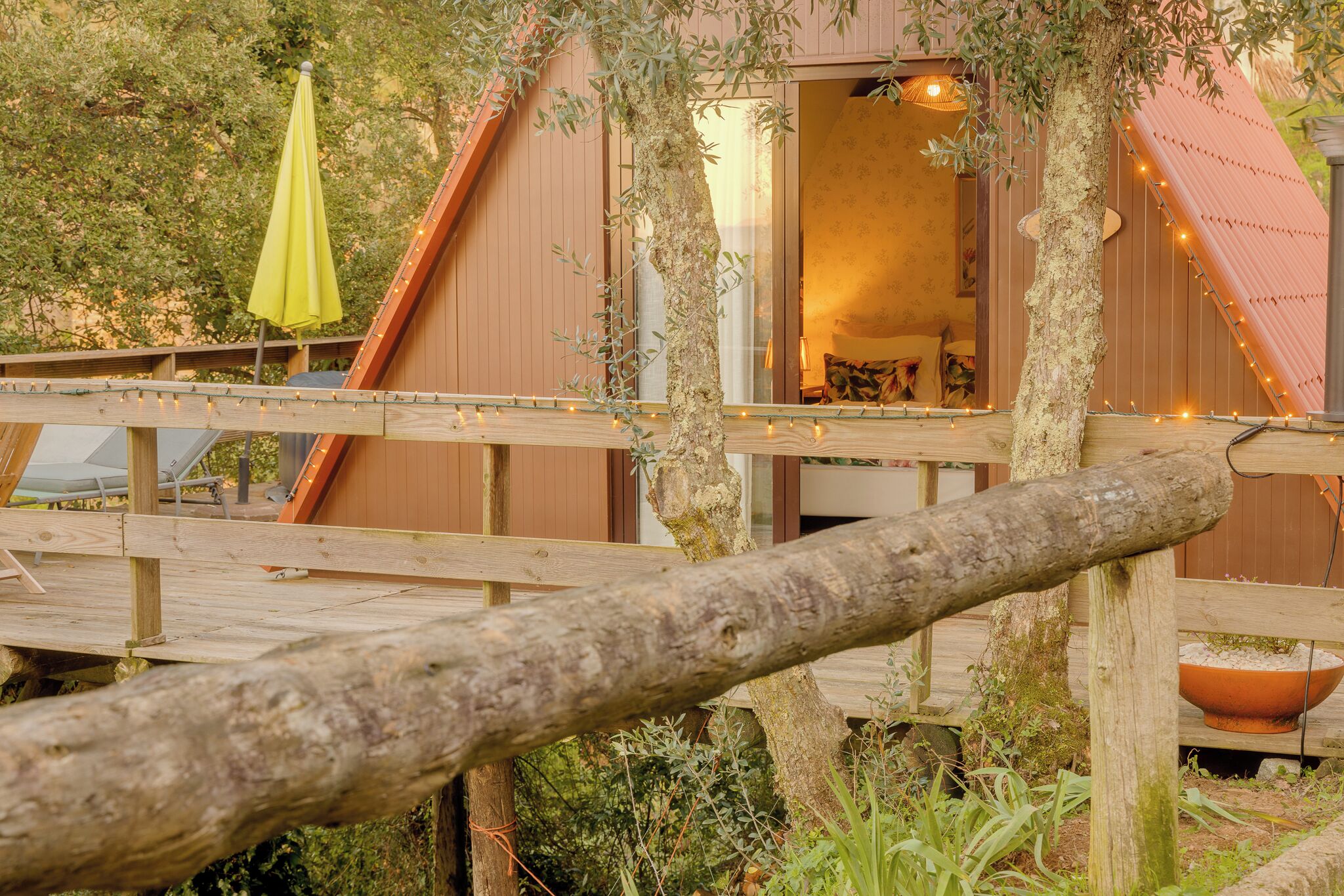 A wooden deck with a rustic tree trunk in the foreground, a wooden building with a window, and a yellow umbrella.