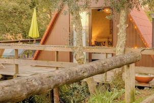 Una terraza de madera con un tronco de árbol rústico en primer plano, un edificio de madera con una ventana y un paraguas amarillo.