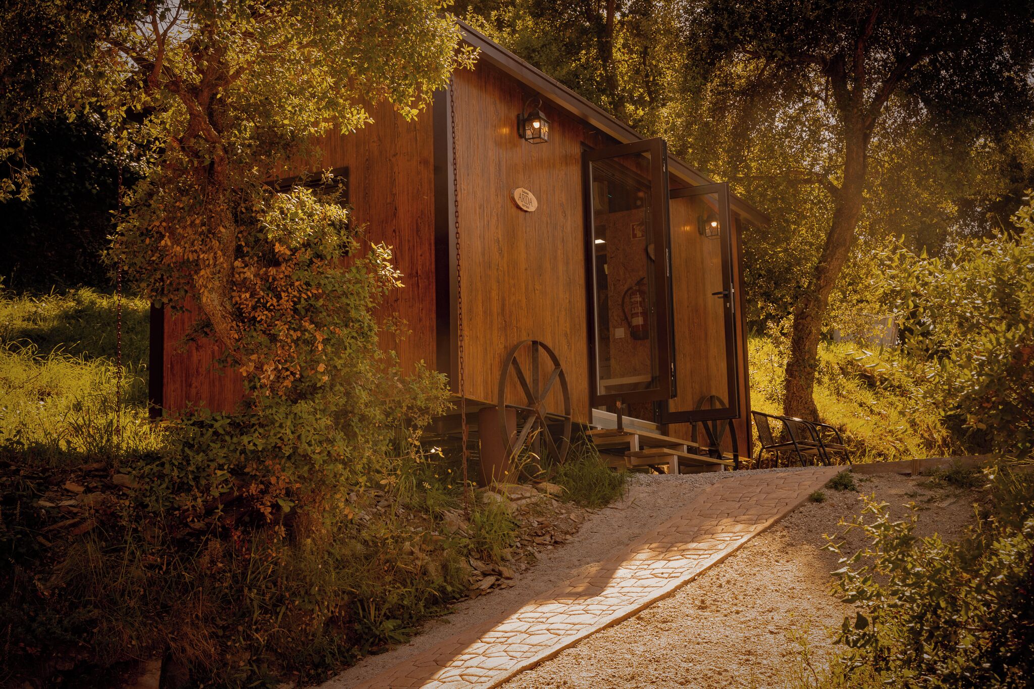 A wooden cabin with a wooden door and a window, surrounded by trees and a gravel path.