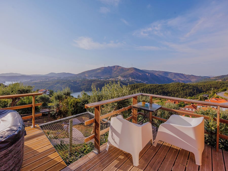 Una terraza de madera con sillas blancas y una mesa, con vista a un paisaje montañoso con aerogeneradores.
