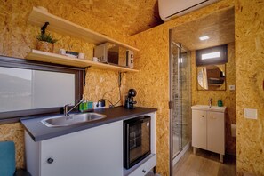 A compact kitchen with a sink, microwave, and oven, a small shelf with a plant and a coffee maker, and a glass shower area.
