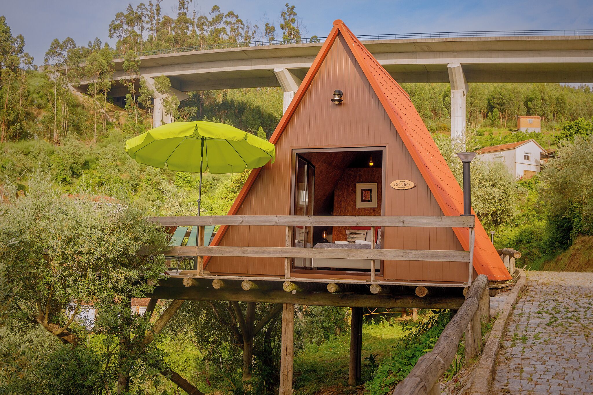 A wooden cabin with a balcony, situated on stilts in a lush green area, with a bridge in the background.