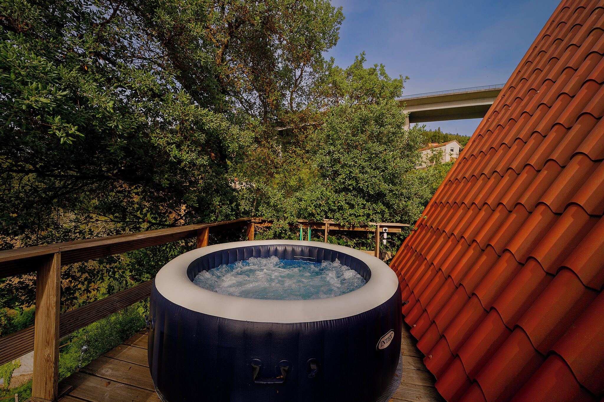 An outdoor hot tub on a wooden deck with a wooden railing, surrounded by greenery and a red-tiled roof.