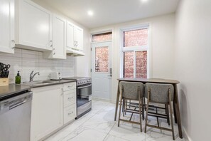 A kitchen with white cabinets, a stainless steel stove, a sink, and a window with a brick wall view. - Warm & Inviting Montréal Stay (Montreal)