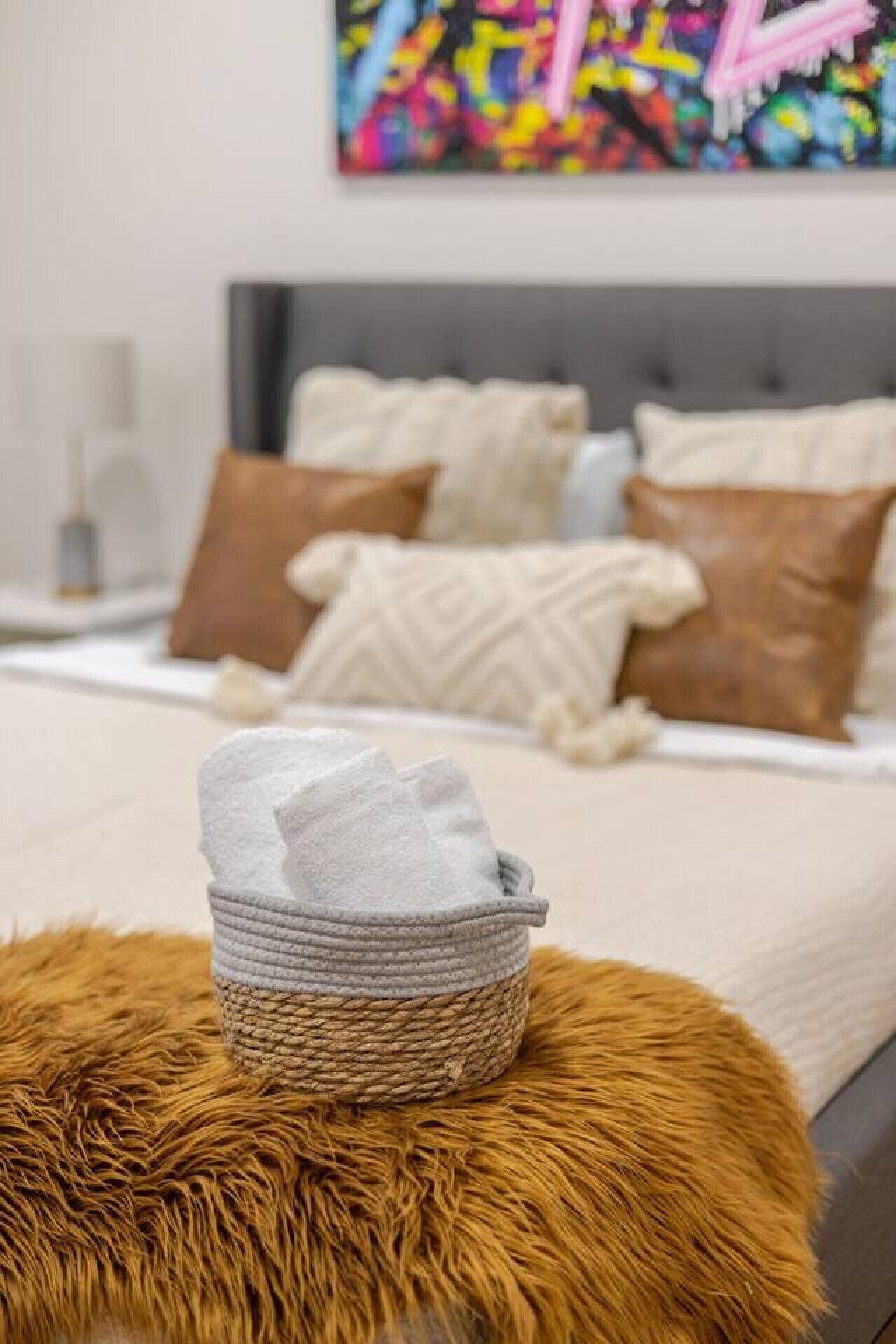 Ein ordentlich bezogenes Bett mit grauer Decke, ein Korb gefalteter Handtücher und ein flauschiger brauner Hocker in einem Hotelzimmer.