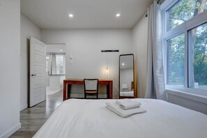 A bedroom with a bed, a desk, a chair, a mirror, and a window with a view of trees. - Cozy & Bright | Prime Location (Montreal)