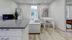 Una cuina moderna amb taulell blanc, zona de menjador amb taula i caires blancs, i una finestra gran amb cortines.