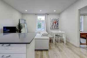 A modern kitchen with a white countertop, a dining area with a white table and chairs, and a large window with curtains. - Cozy & Bright | Prime Location (Montreal)