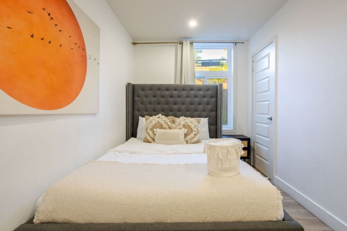 A bedroom with a tufted headboard, a bed with white bedding, a decorative pillow, a bedside basket, and a large abstract painting on the wall.