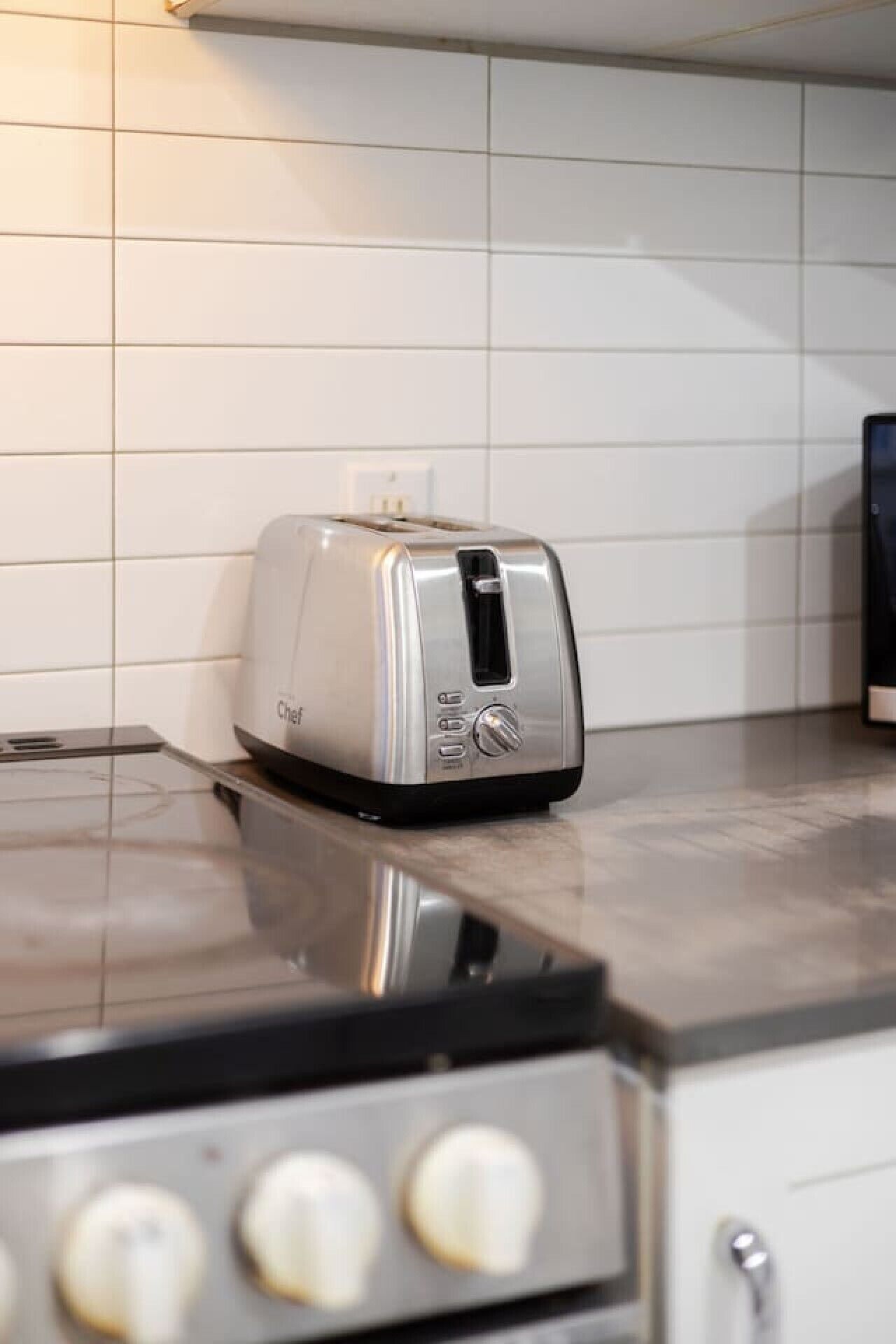 A kitchen with a toaster on the counter, a stove with knobs, and white tiled walls.