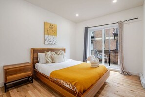 A bedroom with a wooden bed, a nightstand, a window with a view of a brick building, and a painting on the wall. - Prime Downtown Spot | Easy Access (Montreal)