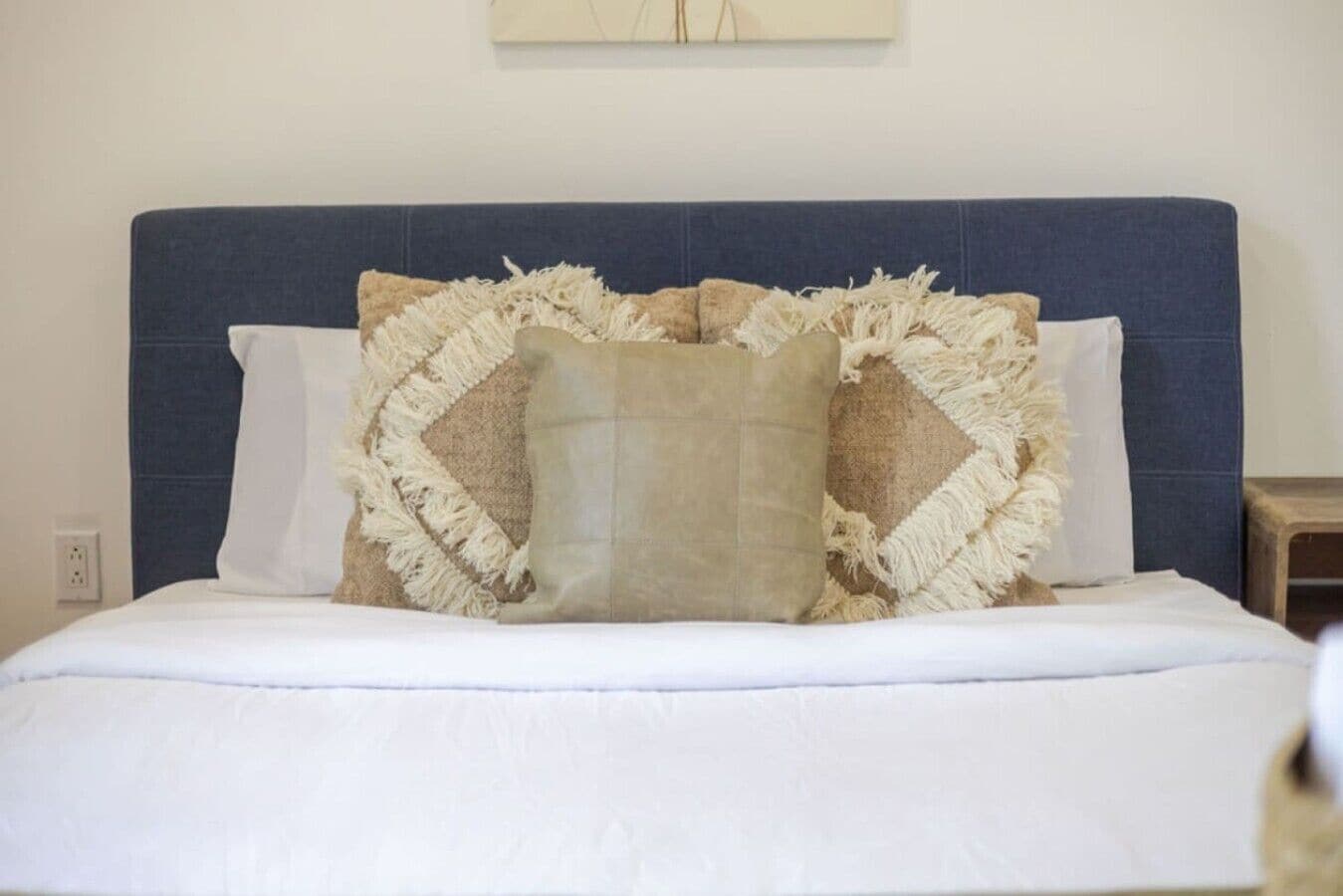 Una cama con cabecera azul, ropa de cama blanca y un cojín decorativo con flecos.