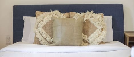 Una cama con cabecera azul, ropa de cama blanca y un cojín decorativo con flecos.