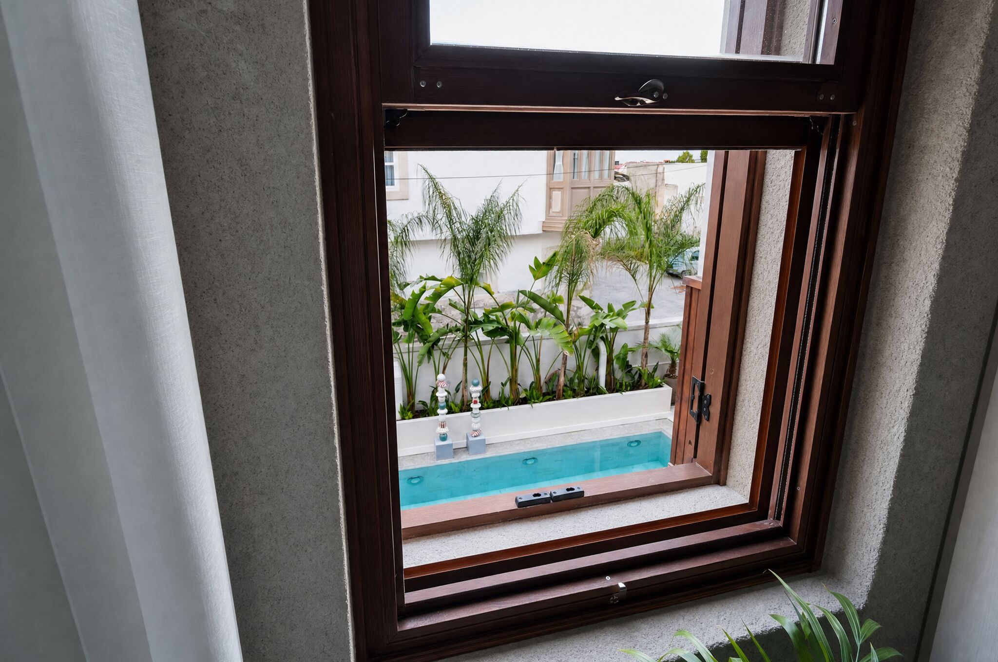 Une fenêtre donnant sur une piscine, des plantes et des bâtiments.