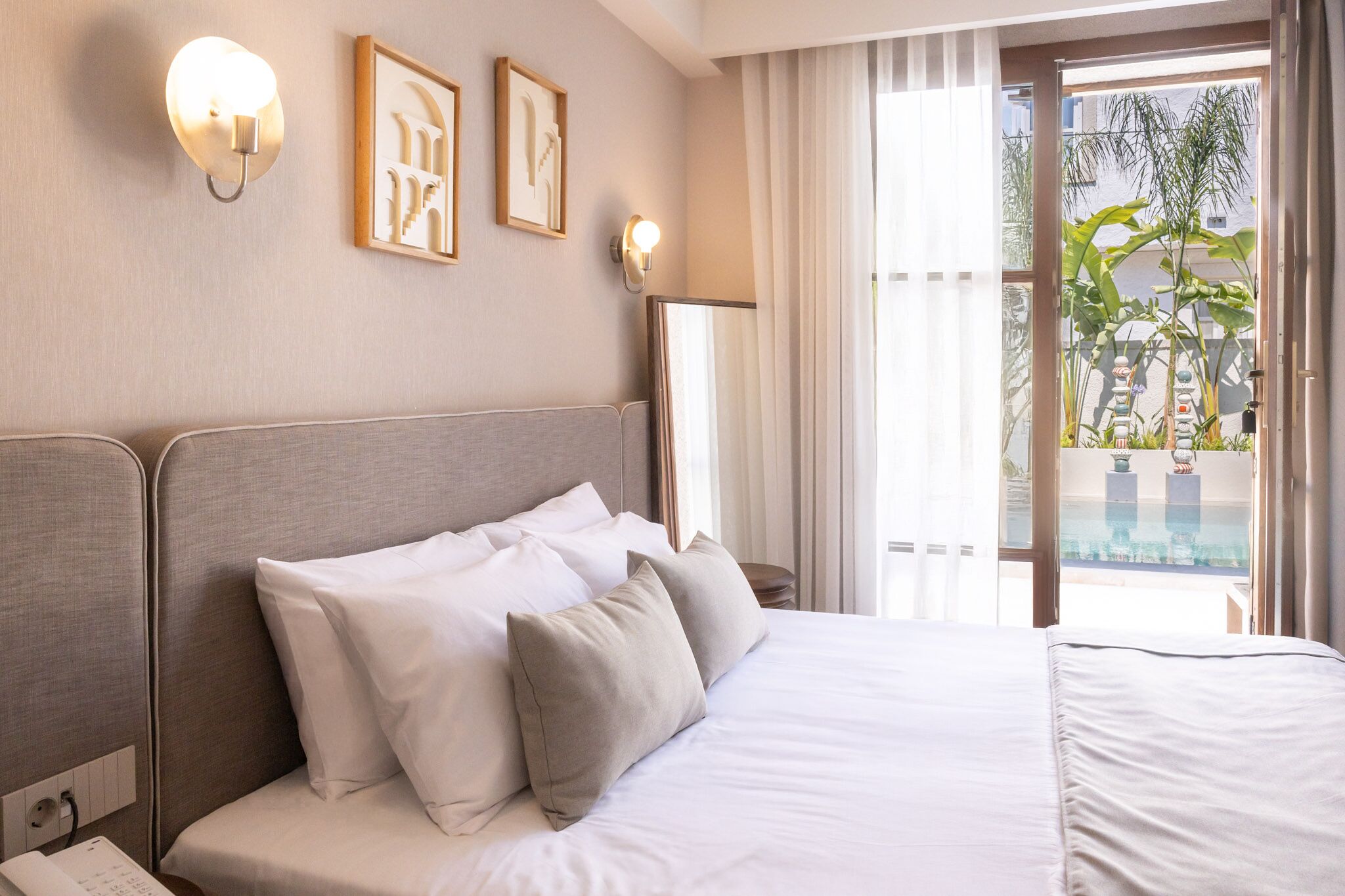 A hotel room with a bed, bedside table, lamp, and a view of a balcony with plants.