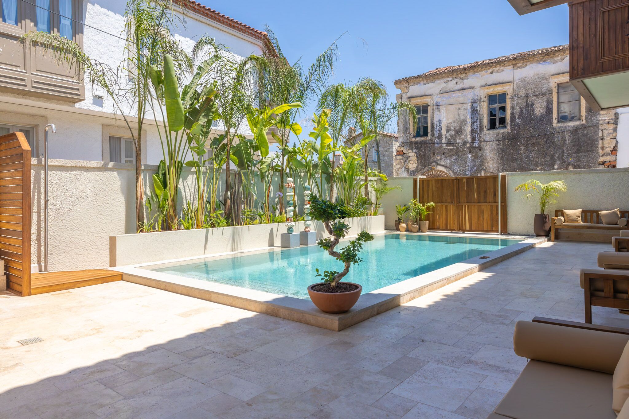 Une piscine agrémentée d’un bonsaï en pot, de mobilier d’extérieur et d’un bâtiment à la façade blanche et au balcon en bois.