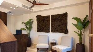 Un área de recepción con escritorio, dos sillas, ventilador de techo y obras de arte en la pared.