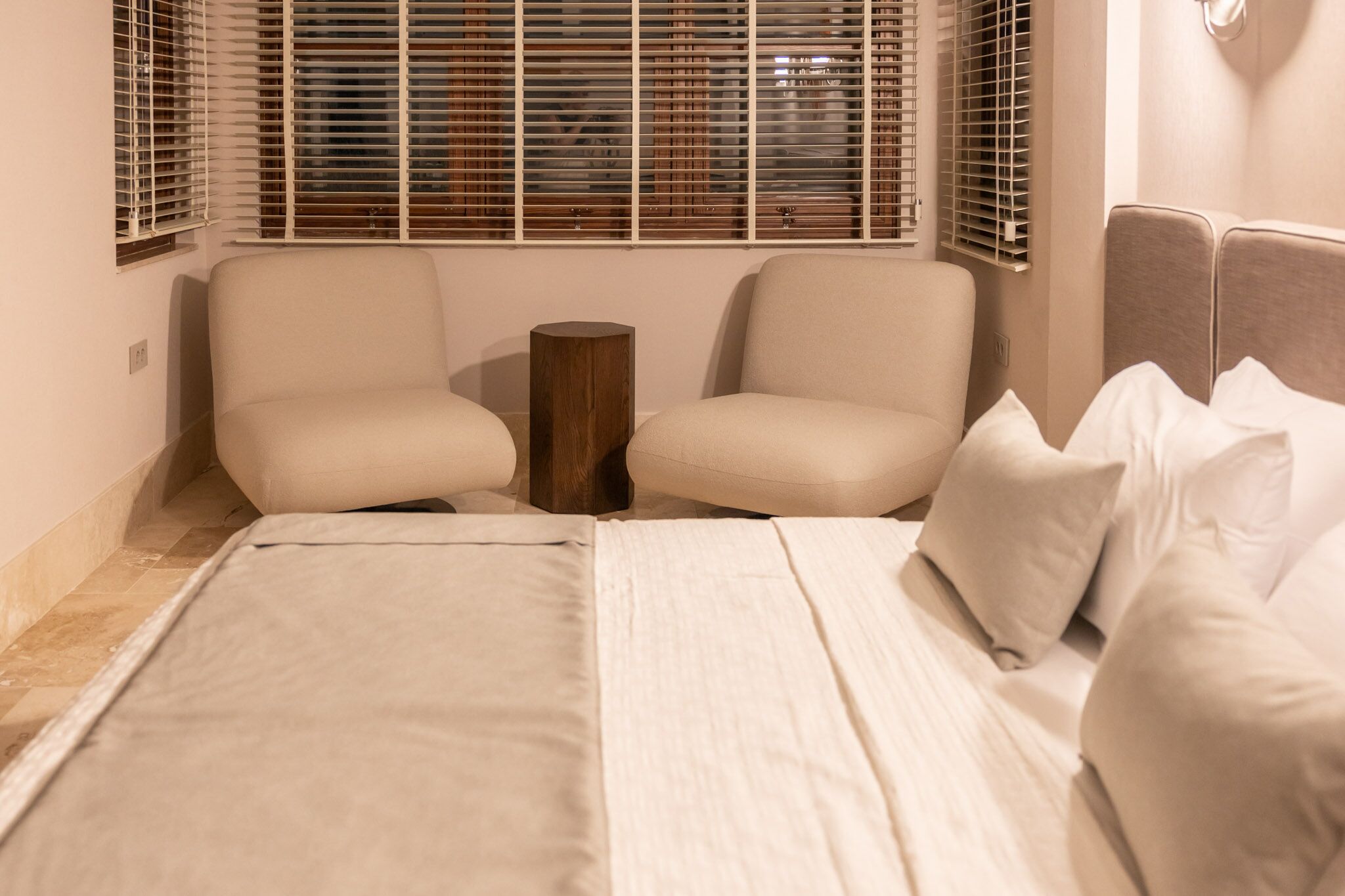 A bedroom with a bed, two chairs, a wooden stool, and a window with blinds.