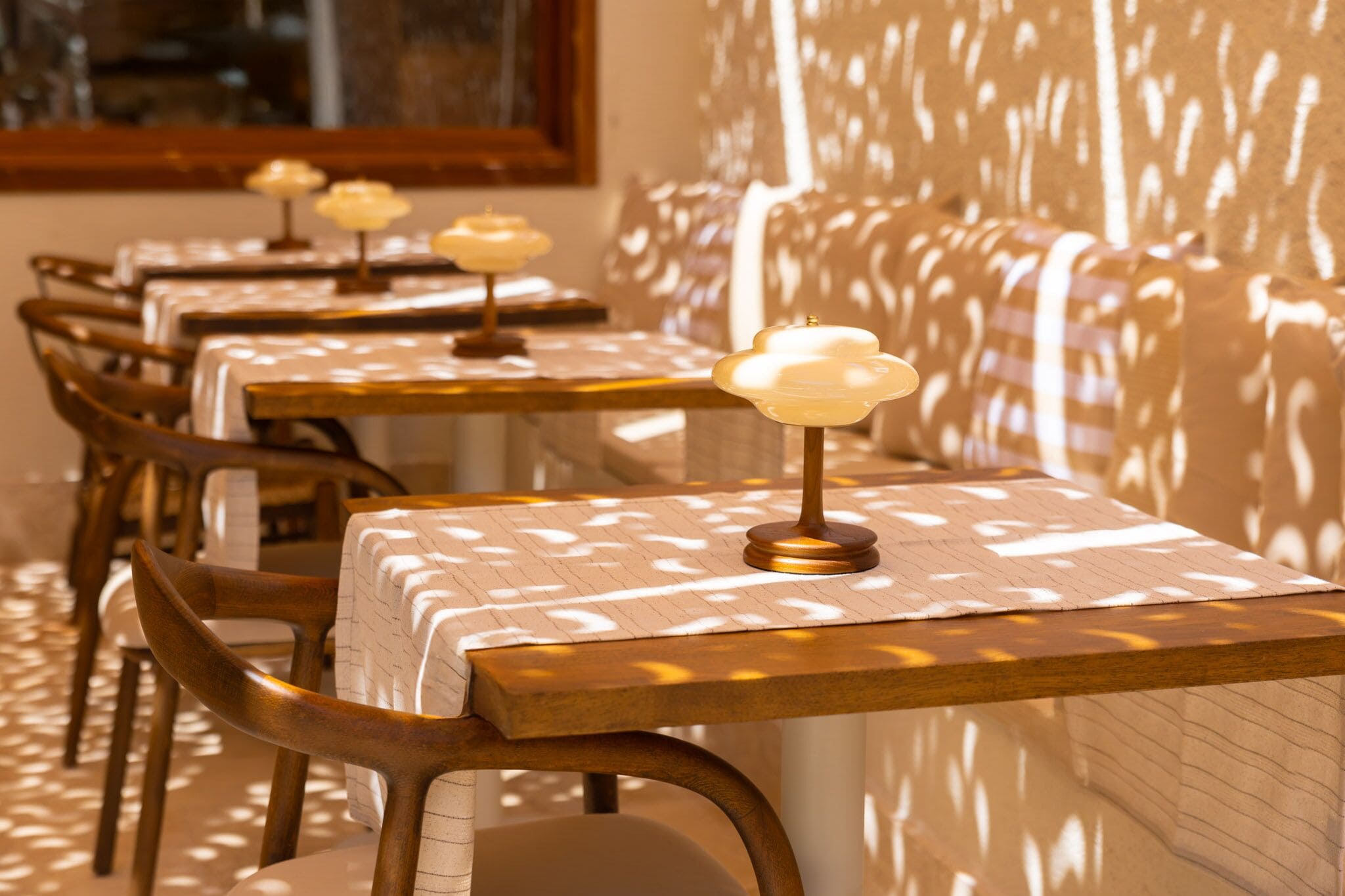 Une rangée de tables recouvertes de nappes à motifs, surmontées de lampes ressemblant à des bougies, des chaises en bois et un mur orné d’un jeu d’ombres.