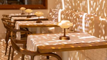 Une rangée de tables recouvertes de nappes à motifs, surmontées de lampes ressemblant à des bougies, des chaises en bois et un mur orné d’un jeu d’ombres.