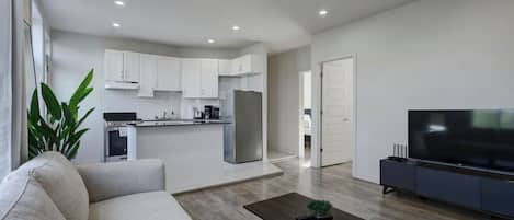 A modern living room with a sofa, coffee table, TV, and a kitchen area with cabinets and appliances.