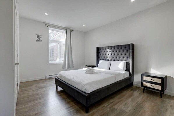A modern bedroom with a large bed, a nightstand, a window with curtains, and a sign on the wall.