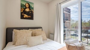 A bedroom with a large bed, a painting on the wall, a window with a view of buildings, and a basket on the floor.
