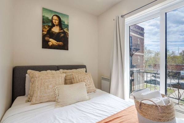 A bedroom with a large bed, a painting on the wall, a window with a view of buildings, and a basket on the floor.