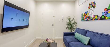 Una sala de estar con un sofá azul, una mesa de centro con un jarrón, un televisor de pantalla plana en la pared y una puerta.