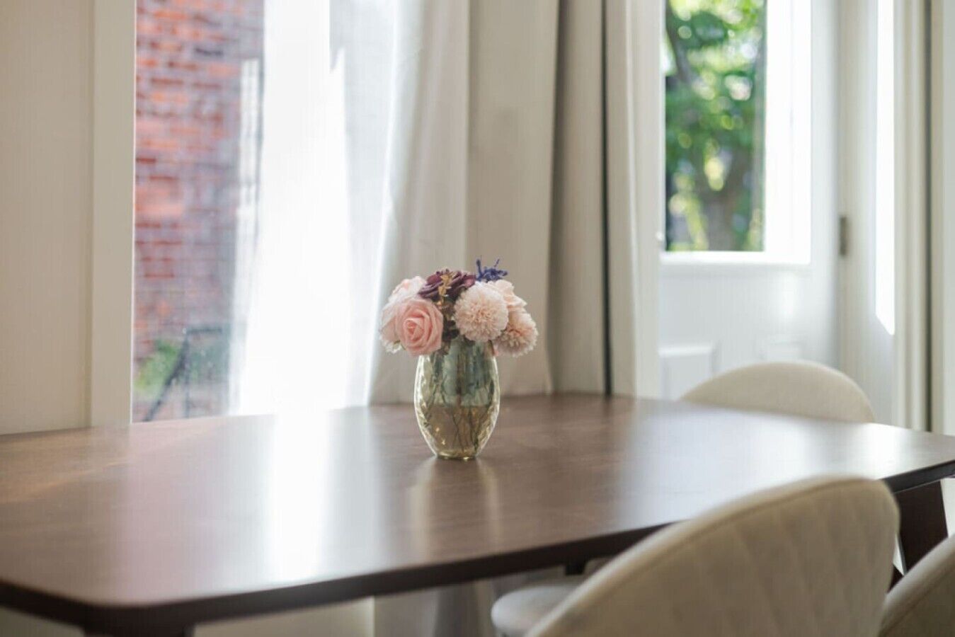 Ein Esstisch mit Blumen in einer Vase, ein Stuhl und ein Fenster mit Vorhängen.