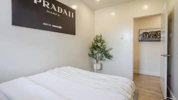 Ein Schlafzimmer mit einem Bett, einem an der Wand befestigten Schild mit der Aufschrift 'PRADA MARFA 1837 MI', einer Topfpflanze und einer Tür zu einem anderen Raum.