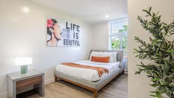 Une chambre avec un lit, une table de chevet avec une lampe, une œuvre d’art murale et une fenêtre donnant sur un paysage verdoyant.