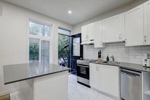 A modern kitchen with white cabinets, a black stove, and a window overlooking a green area. - Stylish Stay in Montréal (Montreal)
