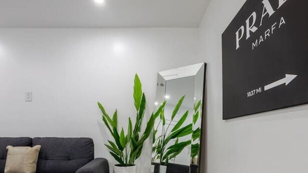 A modern living room with a dark sofa, a potted plant, and a wall-mounted sign reading 'PRADA MARFA 1837 MI'. - Beautiful Stay in MTL (Montreal)