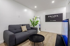 A modern living room with a grey sofa, a small black table, and a TV. A large black sign with 'PRADA MARFA' is on the wall.