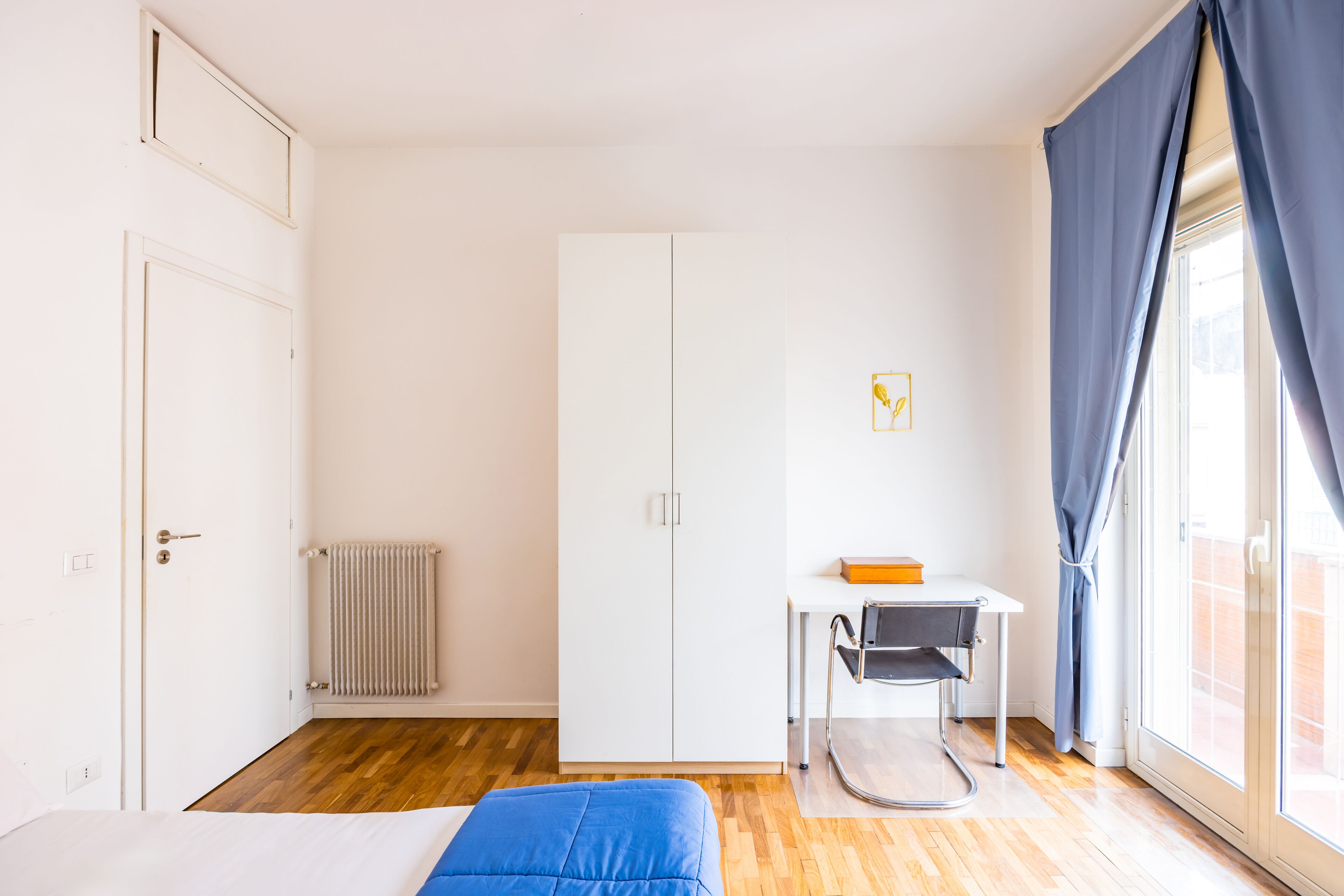 Une pièce comprenant un lit, un bureau, un radiateur et une fenêtre avec des rideaux.