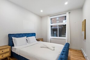 A bedroom with a blue upholstered headboard, white bedding, a wooden nightstand, and a window with a view of a brick building. - Perfect Stay in Montréal (Montreal)