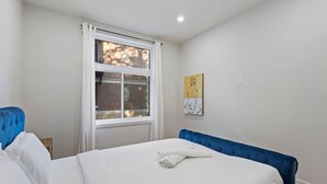 Une chambre avec un grand lit, une fenêtre avec des rideaux, une œuvre d’art accrochée au mur et un mur de briques apparent à l’extérieur.