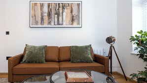 Ett vardagsrum med en brun soffa, två gröna kuddar, ett soffbord i glas, en inramad abstrakt målning, en golvlampa med stativ och en krukväxt.