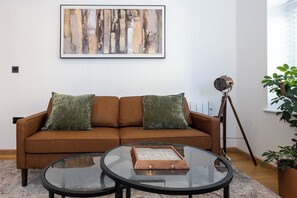 A living room with a brown sofa, two green cushions, a glass coffee table, a framed abstract painting, a tripod floor lamp, and a potted plant. - Modern City Centre 1BR | Walk to Leeds Station (Leeds)