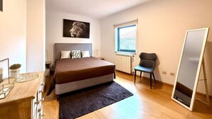 Un dormitorio con una cama, una silla, un radiador, un ventanal y un espejo.