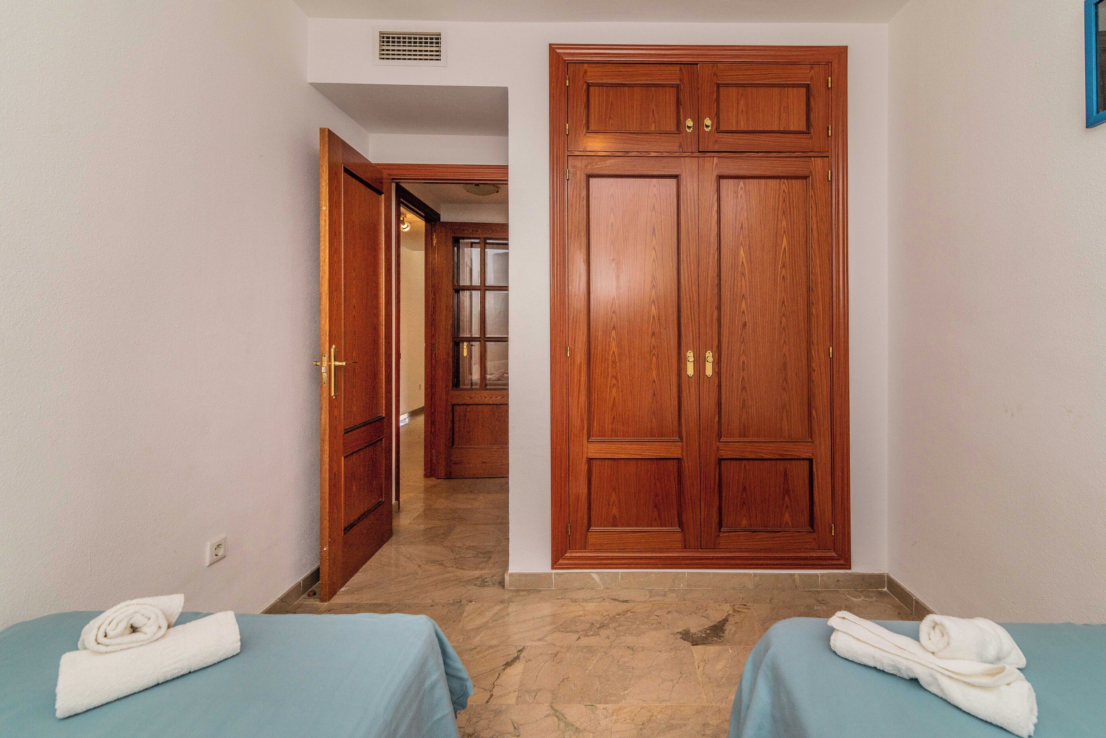 Ein Hotelzimmer mit einem großen Holzschrank, zwei Betten mit blauen Bettlaken und einem durch eine offene Tür einsehbaren Badezimmer.