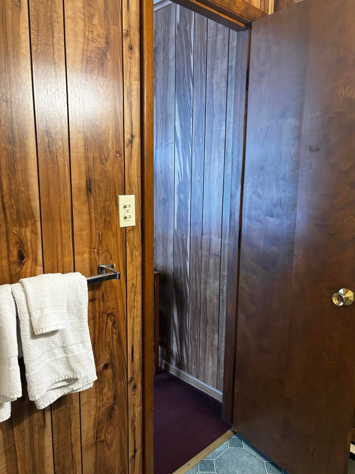 Una puerta de madera con una toalla colgada en un perchero, que da a un cuarto con alfombra morada y piso de madera.