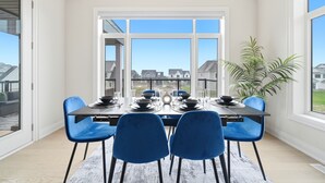 Ein Essbereich mit Tisch für vier Personen, blauen Stühlen, einem Blumentopf und großen Fenstern mit Blick auf angrenzende Häuser.